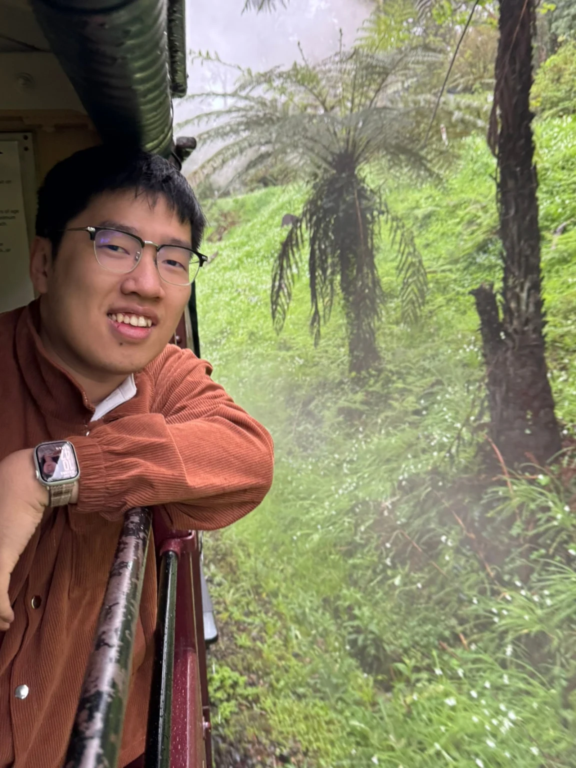 Guian Fang leaning out of a steam train window, smiling, with misty rainforest behind